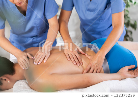 Hands of two masseuses providing four-hand back massage to man 122375259