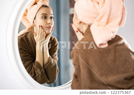 Young woman in bathrobe and towel wrapped around head, looking at mirror 122375367