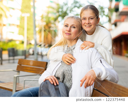 Smiling mom with her teenage daughter sitting on a bench in the center of european city 122375448