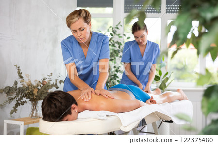 Two female masseuses perform stroking and rubbing of muscles of back and shoulders to guy client Two female masseuses perform stroking and rubbing of muscles of back and shoulders to guy client 122375480