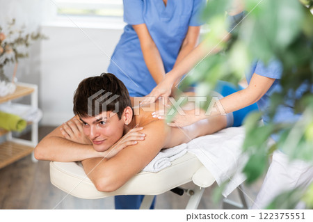 Two woman massage therapist massaging relaxing brunette guy at spa Two woman massage therapist massaging relaxing brunette guy at spa 122375551