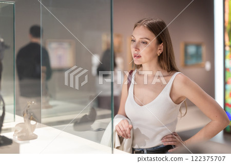 Young female visitor stands near glass display case and examines valuable exhibit 122375707