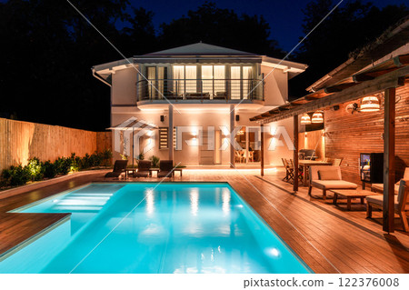 Luxury modern house with illuminated swimming pool and outdoor lounge area at night. Elegant backyard featuring wooden deck and cozy lighting 122376008