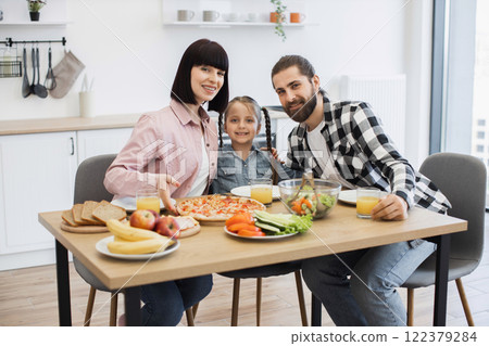 Caucasian family with father, mother, and daughter enjoying pizza and fruit for breakfast at home. Happy morning meal in bright kitchen setting with fresh produce and orange juice. 122379284