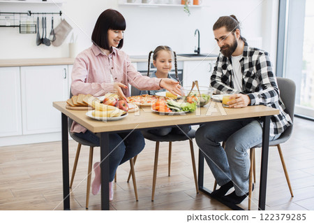Caucasian family with parents and young daughter enjoying breakfast with pizza, fruit, and salad at home kitchen table. Warm family bonding and morning routine. 122379285
