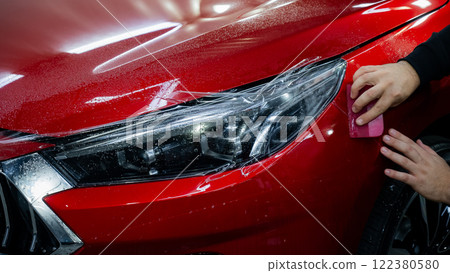 The master applies vinyl film to the headlight of a red car. Closeup view on worker detailer hand smoothing with a scraper protective film. The master applies vinyl film to the headlight of a red car. Closeup view on worker detailer hand smoothing with a scraper protective film. 122380580