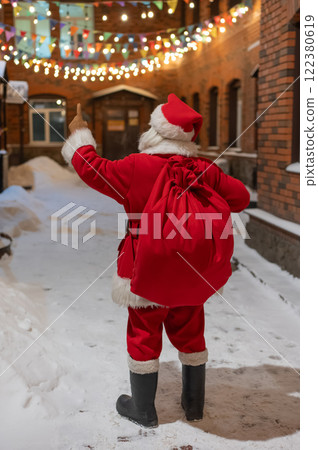 Rear view of santa claus carrying sack of gifts along decorated street. Rear view of santa claus carrying sack of gifts along decorated street. 122380619