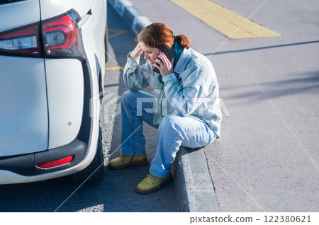 A Caucasian woman sits by a car wheel and calls for help on the phone. 122380621