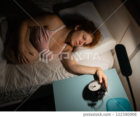 A pregnant woman lies in bed and holds an alarm clock.  122380906