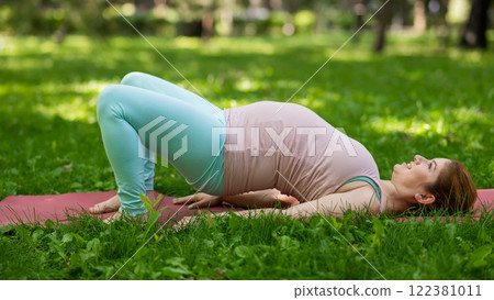 Pregnant caucasian woman doing yoga in the park. Prenatal shoulder bridge exercise.  122381011