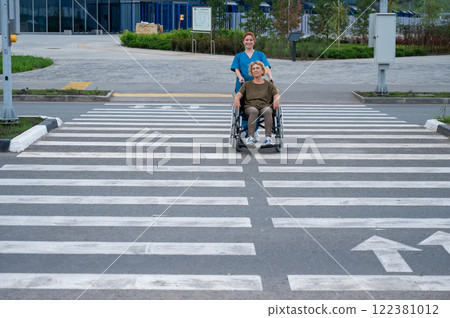 Red-haired nurse pushing an elderly woman in a wheelchair across the road. Red-haired nurse pushing an elderly woman in a wheelchair across the road. 122381012