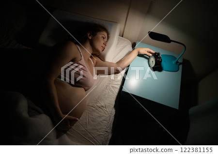 A pregnant woman lies in bed and holds an alarm clock.  122381155