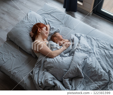 Top view of a red-haired Caucasian woman lying in bed with her baby son. Vertical photo. Top view of a red-haired Caucasian woman lying in bed with her baby son. Vertical photo. 122381399
