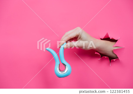 Female hand sticking out of pink paper background with blue curved vibrator sex toy. Copy space. Female hand sticking out of pink paper background with blue curved vibrator sex toy. Copy space. 122381455