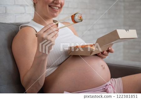 A pregnant woman sits on the sofa and eats rolls from a box. Food delivery.  122381851