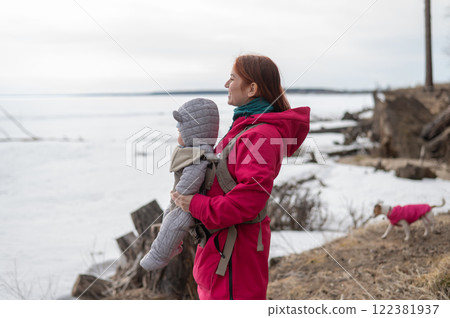 Caucasian red-haired woman walks with her son in an ergo backpack in nature in winter. Caucasian red-haired woman walks with her son in an ergo backpack in nature in winter. 122381937