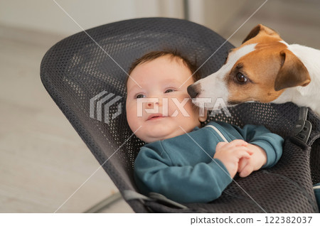 A dog licks the face of a cute three-month-old boy dressed in blue overalls in a baby lounger.  122382037