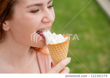 Young beautiful red-haired woman smiling with braces and eating an ice cream cone outdoors. Young beautiful red-haired woman smiling with braces and eating an ice cream cone outdoors. 122382178