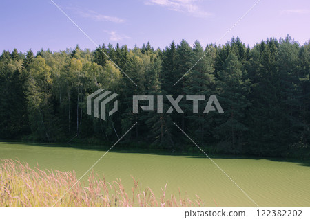 Picturesque view of a forest lake with clear water reflecting green trees and blue sky. The shore is covered with dense coniferous and deciduous trees. Film photography effect and grunge effect 122382202