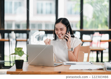 A joyful young woman on a phone call, celebrating success while working on her laptop in a bright, modern office space. 122383466