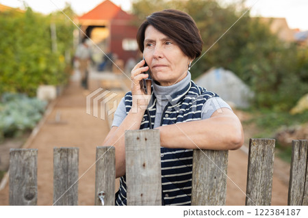 Mature woman standing relaxed near fence talking on phone in backyard smiling Mature woman standing relaxed near fence talking on phone in backyard smiling 122384187