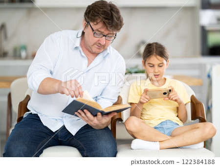 Father reading book, daughter using phone Father reading book, daughter using phone 122384188
