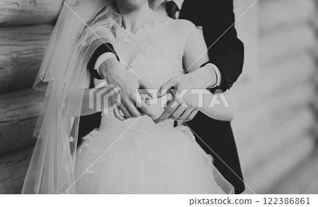 Groom hugs bride in wedding dress from behind, hands show heart. Black and white photo 122386861