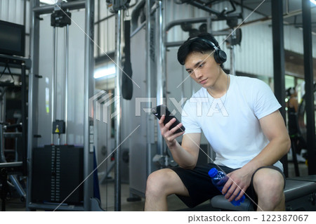 Young sportsman wearing headphones sitting on a gym bench and checking his phone 122387067