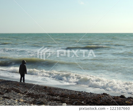 little boy in jacket standing on the shore of cold sea with foamy surf and looking at sea water, lonely child walking along sea stone coast on winter windy day little boy in jacket standing on the shore of cold sea with foamy surf and looking at sea water, lonely child walking along sea stone coast on winter windy day 122387660