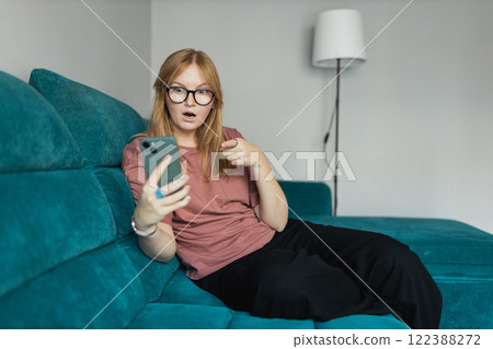 Smiling young woman with smart phone pointing finger a great idea, sitting on a couch at home. Cute beautiful 30s girl chatting by mobile phone 122388272