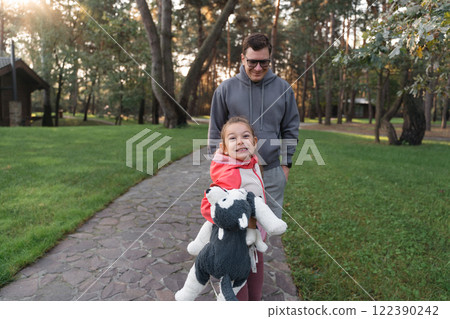 A young girl smiles brightly, holding a stuffed animal, while walking with her father in a green A young girl smiles brightly, holding a stuffed animal, while walking with her father in a green 122390242