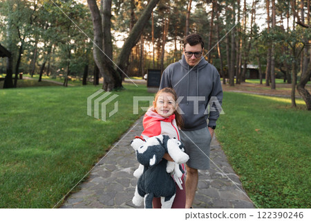 A young girl smiles brightly, holding a stuffed animal, while walking with her father in a green 122390246