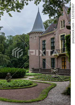 A picturesque view of a historic brick mansion with a round tower, pointed roof, and arched windows, surrounded by lush greenery, manicured gardens, and a peaceful, cloudy sky. A picturesque view of a historic brick mansion with a round tower, pointed roof, and arched windows, surrounded by lush greenery, manicured gardens, and a peaceful, cloudy sky. 122391867