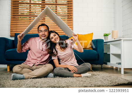 In their new empty apartment an Asian husband and wife bond while crafting a cardboard home roof with their hands. Full-length portrait against a white studio wall a symbol of achievement and joy. 122393951