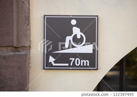 Rectangular accessible entrance sign, featuring wheelchair disability icon and arrow on the wall outdoors. Accessible route for handicapped sign posted on the wall Rectangular accessible entrance sign, featuring wheelchair disability icon and arrow on the wall outdoors. Accessible route for handicapped sign posted on the wall 122397495