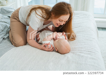 Happy family at home. Mother playing with little toddler child on bed. Mom baby relax playing having fun together. Mother looks at baby with love care. Mom of breast feeding baby rest in living room Happy family at home. Mother playing with little toddler child on bed. Mom baby relax playing having fun together. Mother looks at baby with love care. Mom of breast feeding baby rest in living room 122397734