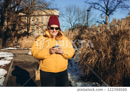 Young stylish 30s woman in sunglasses writes message on smartphone walking on city street. Urban lifestyle concept. Winter time. Charming female wearing stylish outfit chatting online 122398080