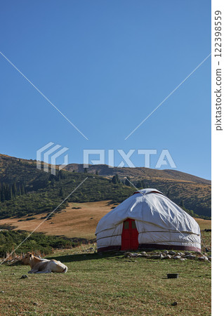 Serene Yurt Positioned Amidst Nature, Complemented by a Vast Grassland Background. cow is lying nearby beautiful, picturesque yurt is set amidst a lush, green landscape, all under vast, clear blue sky 122398559
