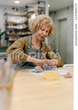 Elderly Caucasian woman sculpts clay bowl at pottery workshop, learning new skill. Senior lady enjoys working with hands, developing creativity and fine motor skills 122400074