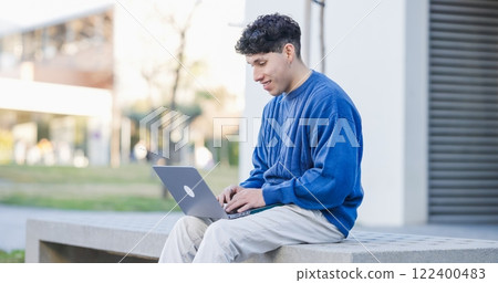 Young hispanic student working on laptop sitting on bench in university campus 122400483