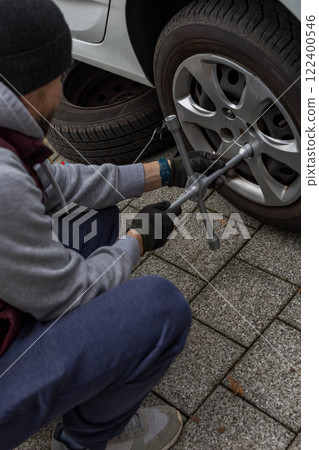 Young man changing a car tire on the road, using a wrench to unscrew the lug nuts Young man changing a car tire on the road, using a wrench to unscrew the lug nuts 122400546