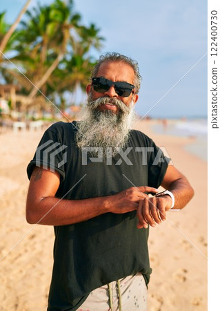 Senior bearded man with sunglasses on sandy beach uses smart watch. Modern grandpa enjoys tech, staying fit by the sea. Active old stylish male uses wearable device in sunny tropical location. Senior bearded man with sunglasses on sandy beach uses smart watch. Modern grandpa enjoys tech, staying fit by the sea. Active old stylish male uses wearable device in sunny tropical location. 122400730
