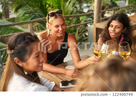 Diverse women friends toasting with wine glasses at restaurant. Social evening out, celebrate friendship at dinner party. Tropical vibes, casual dining, fun, laughter. 122400749