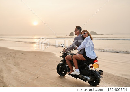 Couple rides motorcycle on beach at sunset. Travel on bike by ocean in warm country. Relaxing vacation, exploring tropical coast, enjoying waves. Adventure seekers on relaxed journey. 122400754