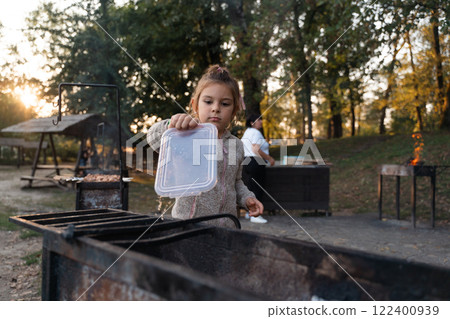 A young girl assists with a barbecue by holding a container over the grill, surrounded by trees and A young girl assists with a barbecue by holding a container over the grill, surrounded by trees and 122400939