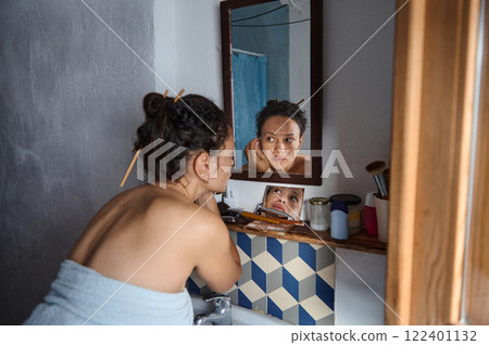 Woman applying makeup while reflecting in a bathroom mirror at home Woman applying makeup while reflecting in a bathroom mirror at home 122401132