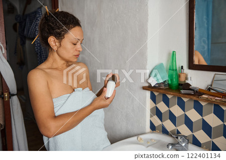 Woman standing in bathroom examining cosmetic bottle wrapped in towel 122401134