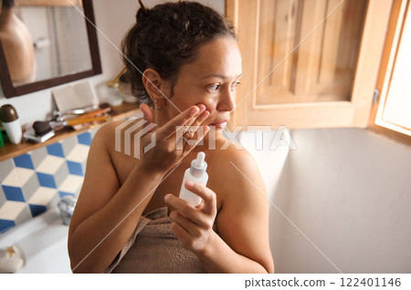 Woman applying skincare product in a well-lit home bathroom setting Woman applying skincare product in a well-lit home bathroom setting 122401146