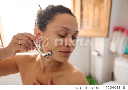 Woman using a roller massager on her face in a bright bathroom Woman using a roller massager on her face in a bright bathroom 122401148