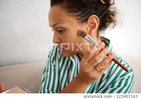 Woman applying makeup with a brush in a relaxed indoor setting Woman applying makeup with a brush in a relaxed indoor setting 122401163